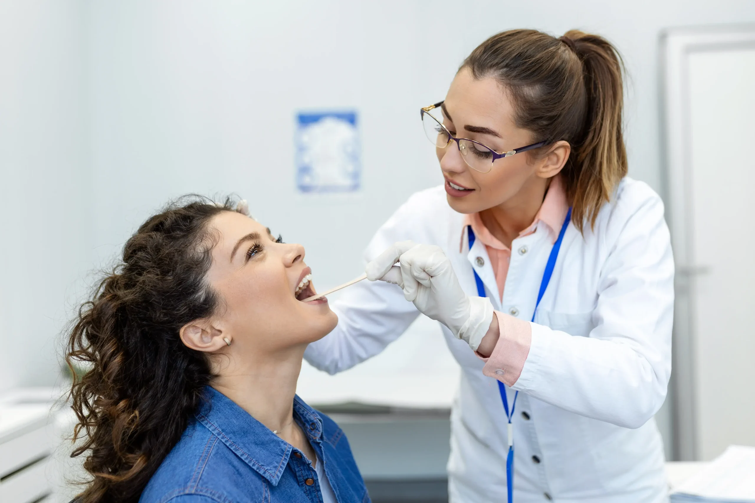 A female patient opens her mouth as an ENT doctor uses an inspection spatula to examine her throat during a routine checkup.